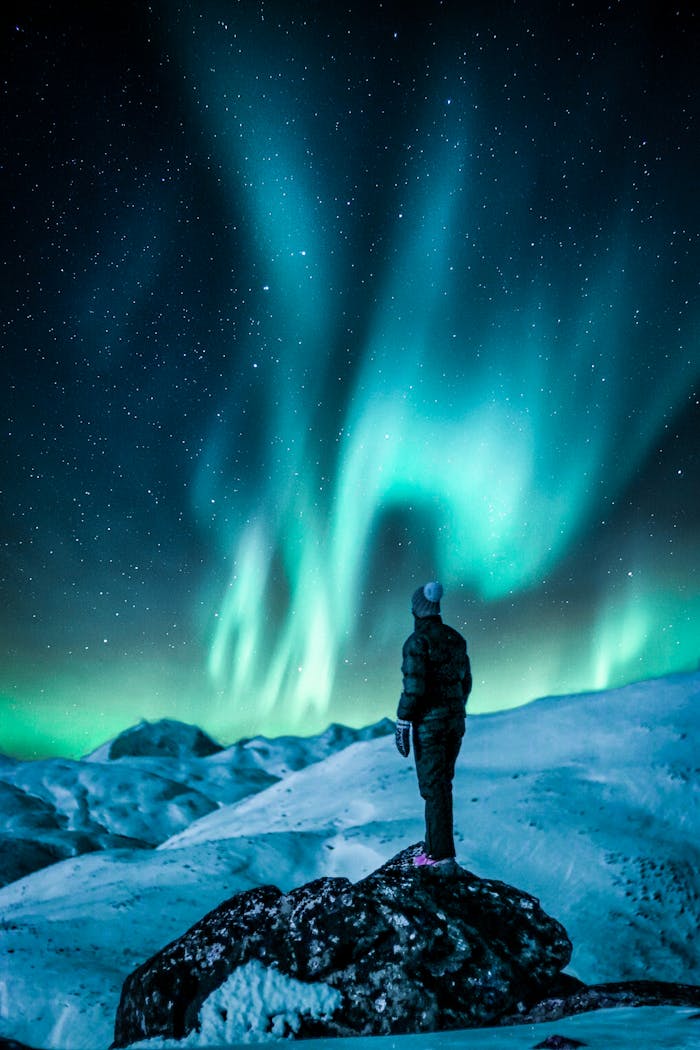 digital-01 A lone figure stands on a snowy slope, gazing at the vibrant Northern Lights under a starry sky.